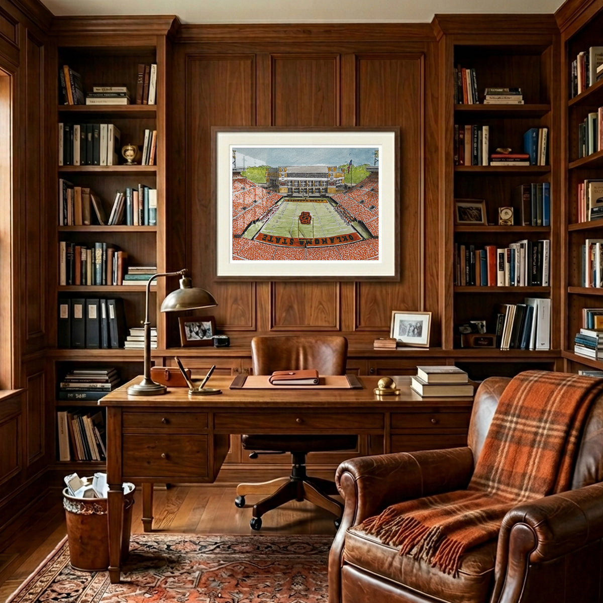 A framed art print depicting Boone Pickens football stadium at Oklahoma State, displayed on a dark wood-paneled wall in a classic study with a desk, leather chair, and bookshelves.