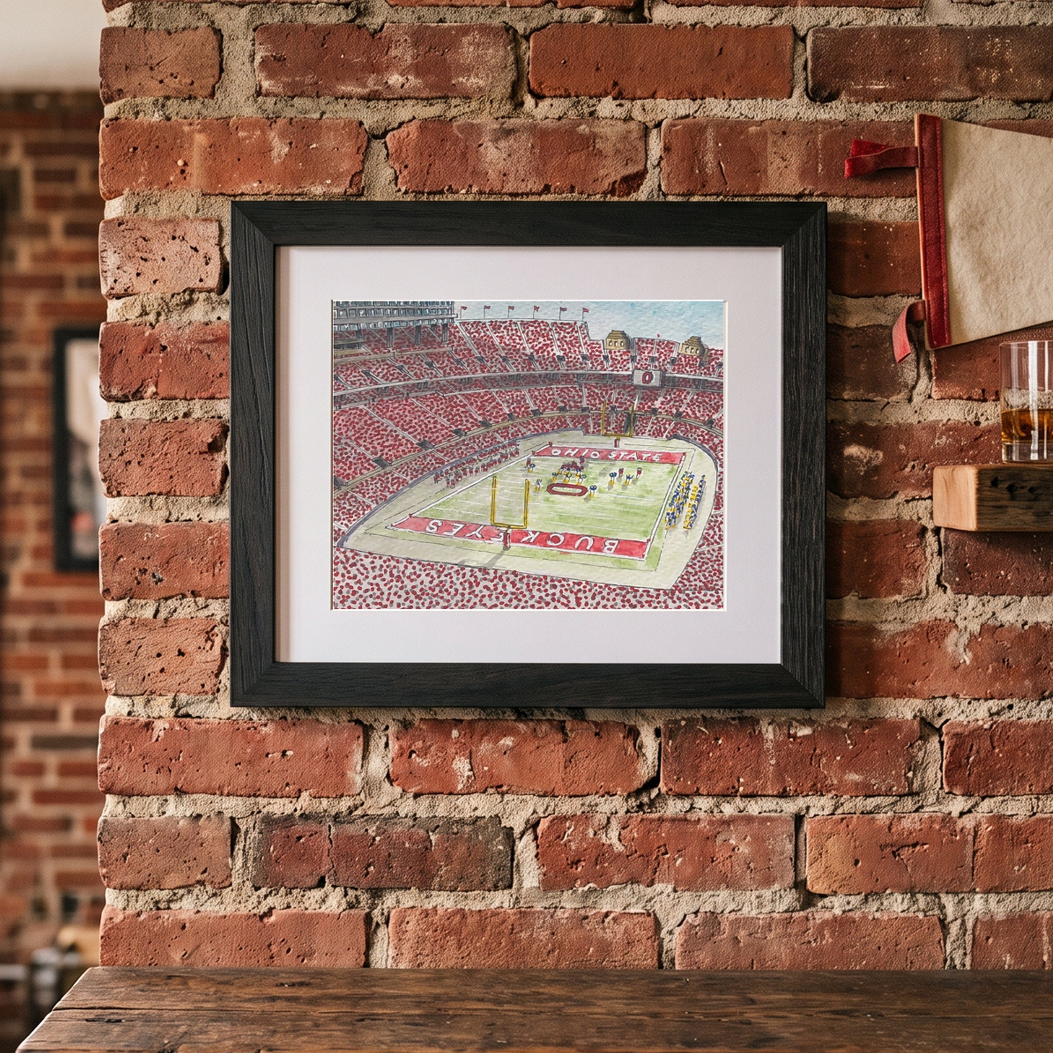 An architectural art print of Ohio State University football stadium mounted on an exposed brick wall above a clean wood shelf.
