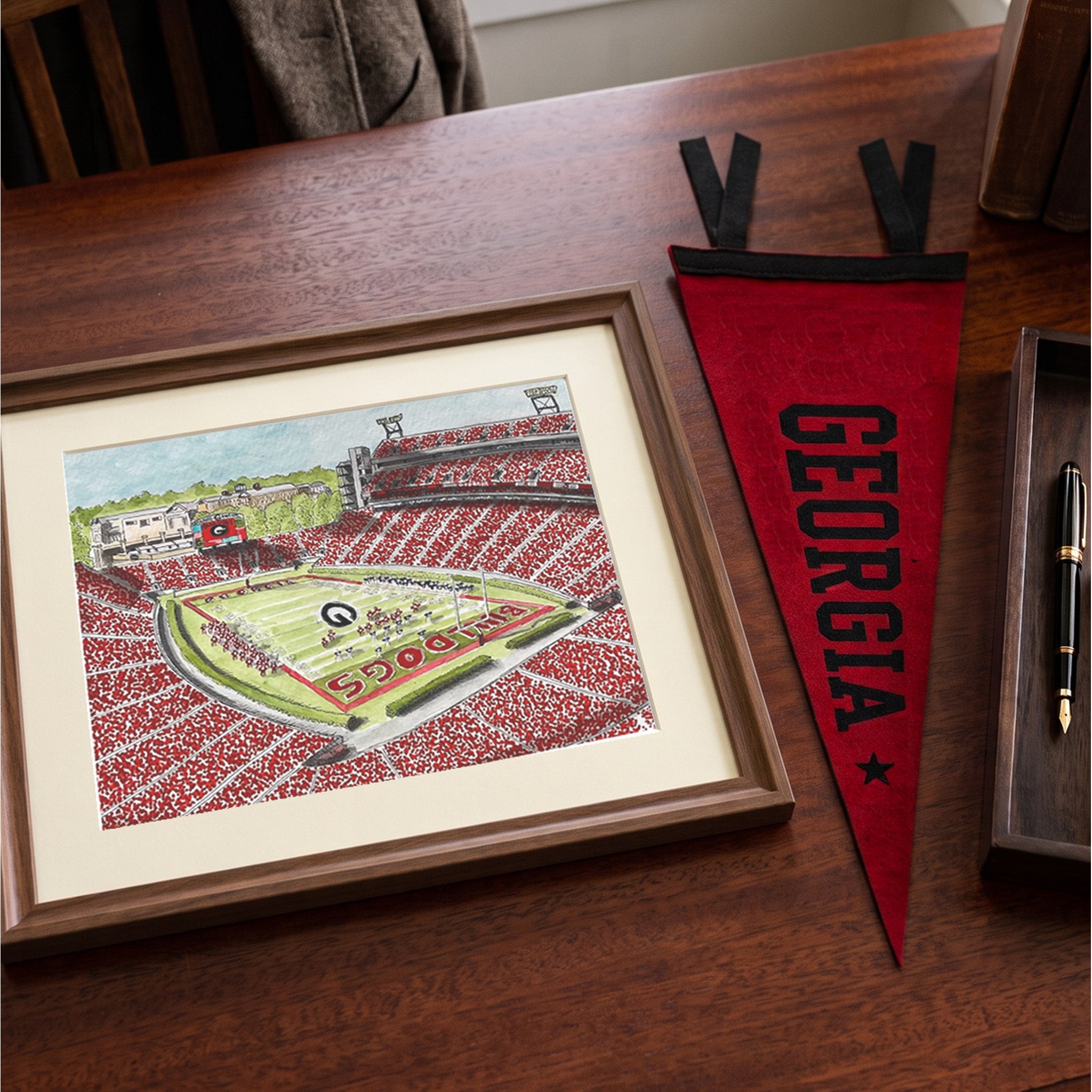 A framed hand-drawn illustration of a football stadium with the word 'GEORGIA' on a red pennant beside it, displayed on a wooden desk next to a pen.