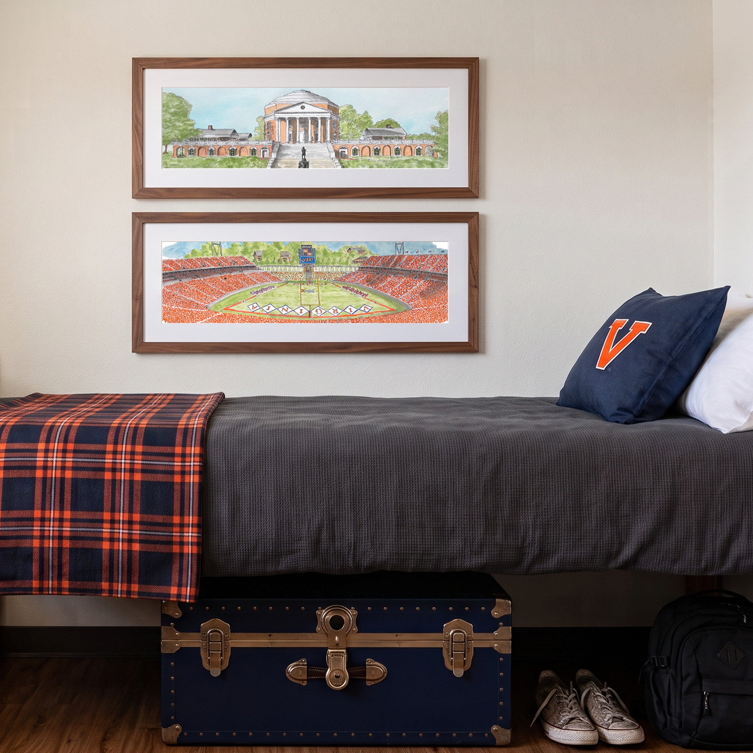 A bedroom scene with two framed art prints depicting a classical building and a football stadium hanging above a bed. The bed has a dark gray textured cover, a navy plaid throw blanket, a navy pillow with an orange letter 'V', and a blue trunk underneath.