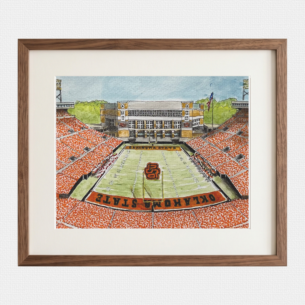 Framed watercolor art print of an Oklahoma State football stadium, featuring a green field, orange stands, and the Oklahoma State logo, presented in a brown wood frame with a white mat.